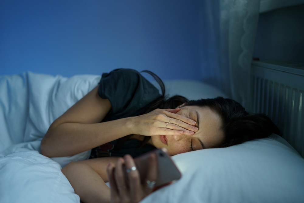 woman looking at phone while lying in bed and rubbing tired eyes