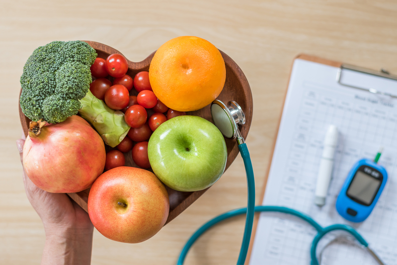 heart-shaped bowl of fruits and vegetables next to stethoscope and blood sugar test device